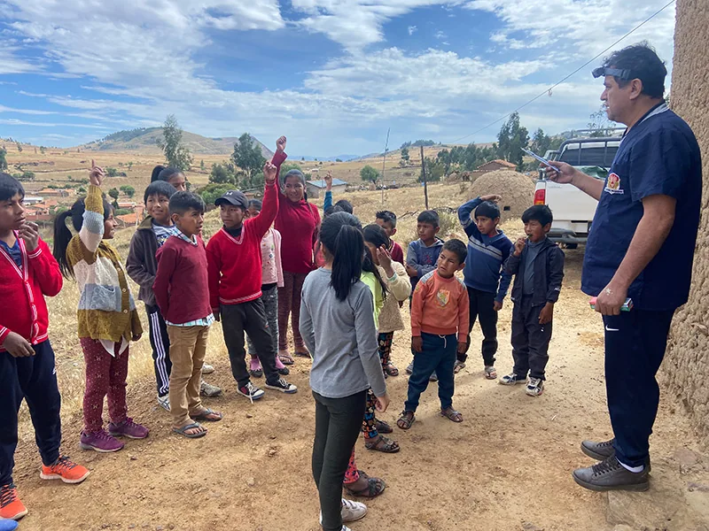 A man in dark scrubs stands on a dirt slope addressing about fifteen children, several with their hands raised.