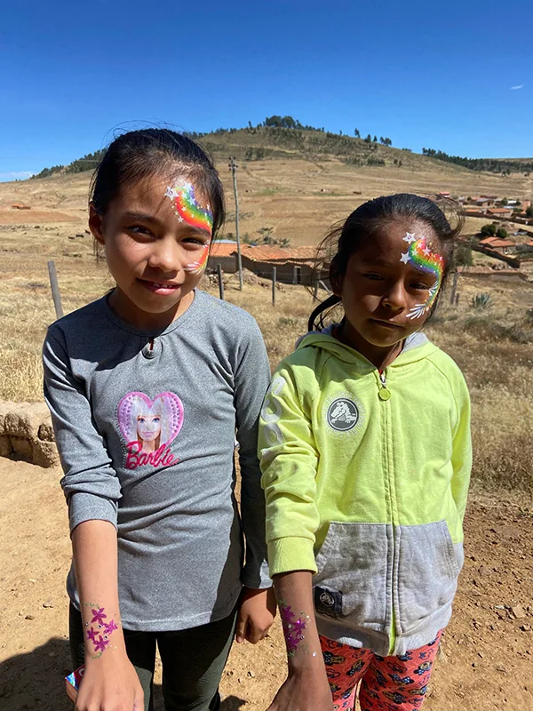 Two girls with rainbow face paint and glitter stars stand together on a hillside above a village of red roofs.